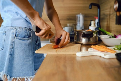 Close-up view of unrecognizable woman slicing fresh carrot lengthwise on wooden board. Pots and