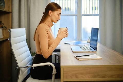 Woman sits at desk in her home office. She holds a cup and looks intently at her laptop screen