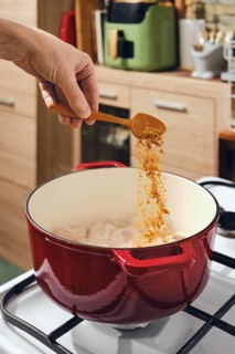Hand adds spice from wooden spoon to cast iron pot on stove. Kitchen has cabinets and counter