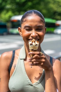 Young woman smiling while holding and eating an ice cream cone outdoors on a city street, conveying