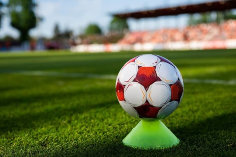 Red and white soccer ball placed on a training cone on a grass pitch with stadium seating in the