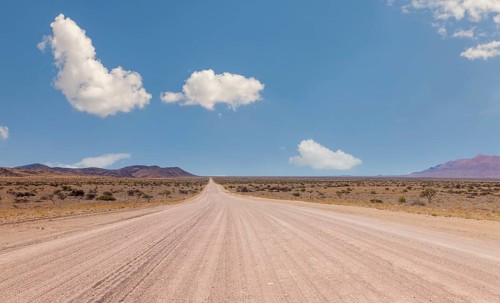 Endless dusty road through dry desert under blue sky with scattered clouds, Namibia