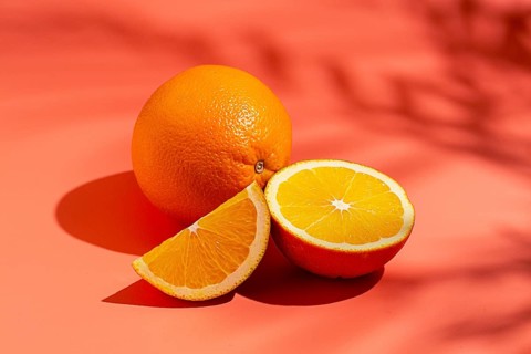 On the coral surface, next to two neatly sliced slices, there is a whole orange. Sunlight casts