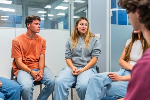 Young adults sitting in a circle, listening to a peer speaking during a group therapy session,