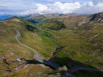 Winding road through green mountain valley under a sky full of clouds, aerial view, near Rânca,