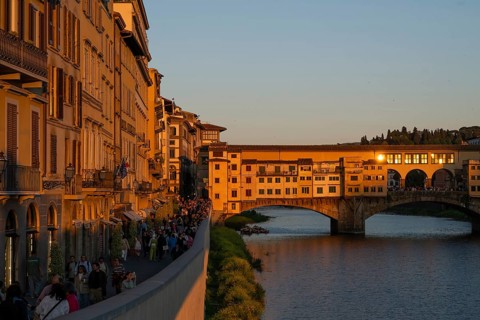 People stroll along the Arno River at sunset, warm evening light falls on the historic Ponte
