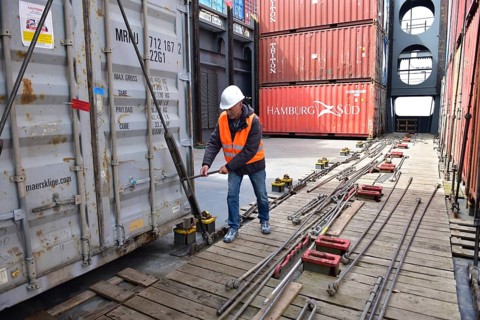 In the port, a man with safety equipment works on stacks of containers on a wooden platform,
