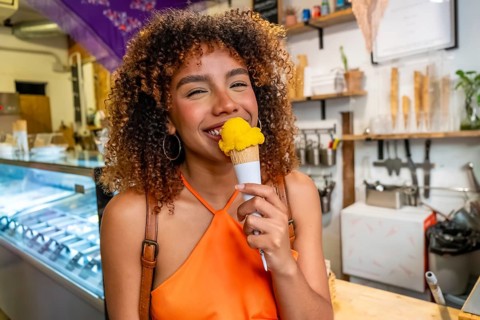 Smiling young woman with curly hair eating a refreshing scoop of yellow ice cream from a waffle