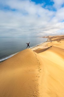 Young woman jumping into the air on a sand dune in front of impressive desert landscape and sea,