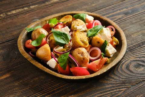 Warm potato and feta salad is mixed with fresh tomatoes and herbs. Dish is served on wooden plate.