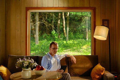 Middle-aged man hiding from heat in wooden cabin outside city sitting on couch and reading