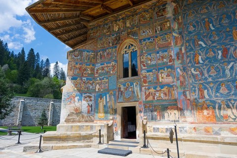 Church wall with colorful frescoes surrounded by trees under a blue sky, exterior view, Saint
