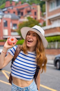 Young smiling woman wearing a hat and striped top holding a pink glazed donut with sprinkles, happy