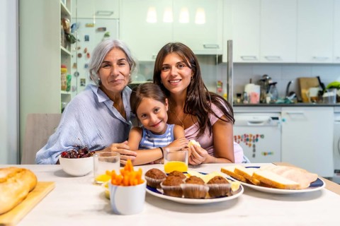 Multi generational women bonding, sharing a meal, and smiling at the camera at a kitchen table,