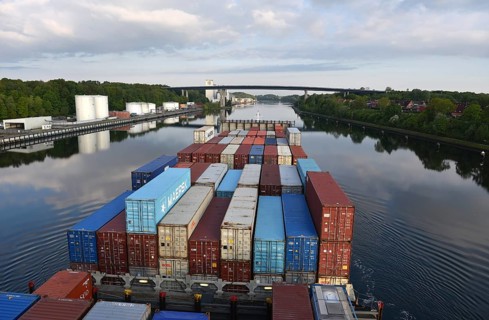 A container ship sails under a bridge on a quiet river, surrounded by trees, container ship sails