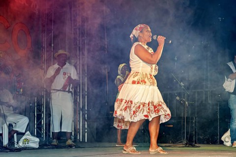 Woman in traditional clothing style sings with fog effects on a stage, a band plays and dances at a