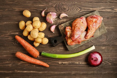 Raw marinated rabbit meat on a cutting board along with potatoes, carrots, garlic, and onions.
