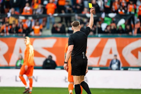 Back view of a soccer referee in a black uniform holding up a yellow card for a foul during a