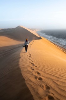 Young woman with camera, sand dunes with wave pattern and sea in evening light, wind blowing sand