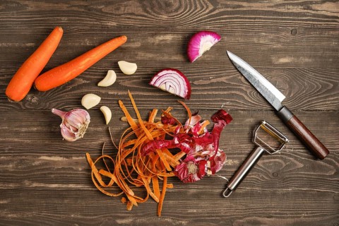 Two carrots, several garlic cloves, and pieces of red onion are laid out on a wooden table. A knife