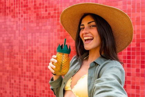 Woman in a straw hat and bikini top laughing while holding a tropical pineapple drink, taking a
