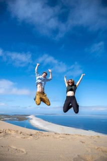 Tourists, young couple jumping in the air, stretching arms in the air, view of high dunes of the