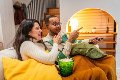 Happy diverse couple relaxing on a couch, holding popcorn, pointing while enjoying an entertaining