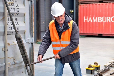 Worker wearing helmet and safety vest uses metal bar while working on a container in port, worker