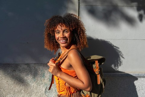 Young woman with natural curly afro hair and backpack walking in sunlit city streets, smiling