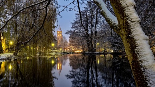 A snow-covered river with illuminated tower and trees in the evening light, reflected in the water,