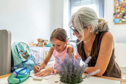 Grandmother is patiently helping her young granddaughter with homework at a desk in a light filled
