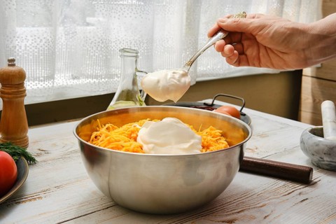A person adds a spoonful of sour cream to a bowl of shredded cheese and chicken meat in a kitchen.