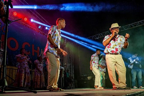 Musicians in floral shirts perform with microphone and instruments at night, a band plays and