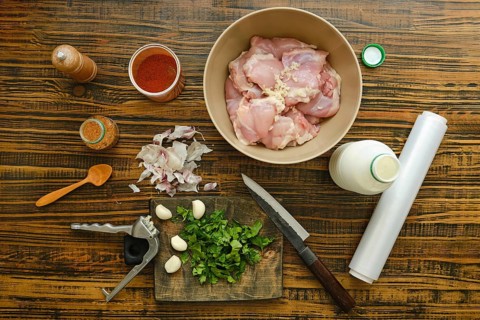 Chicken thighs are in bowl surrounded by spices, herbs, garlic, and yogurt. A knife and cutting