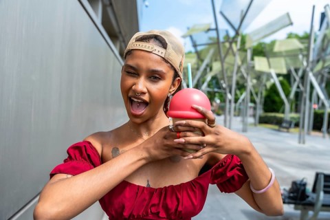 Playful young woman winking at the viewer, holding a fun red capped drink with hands wrapped around