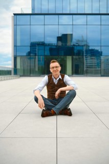 Cheerful middle-aged man sits cross-legged on floor of outdoor terrace. Glass buildings stand tall