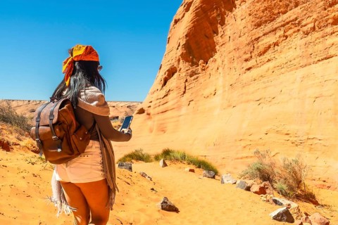 Woman with backpack and smartphone walking a sandy trail toward biidi the arch cave in page,