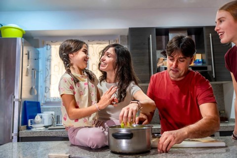 Family including parents and children standing and sitting in a bright kitchen, happily engaging in