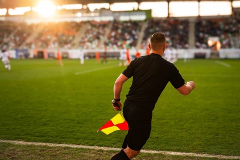 Rear view of a soccer assistant referee in black running with a flag on a pitch at sunset during a