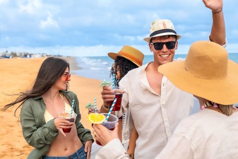 Young friends toast cocktails on a sunny sandy beach at sunset, smiling and celebrating summer