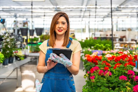 Woman gardener wearing overalls and gloves, smiling while using a digital tablet to manage plant