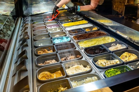 Person wearing gloves scooping vibrant gelato flavors from a refrigerated display case inside an