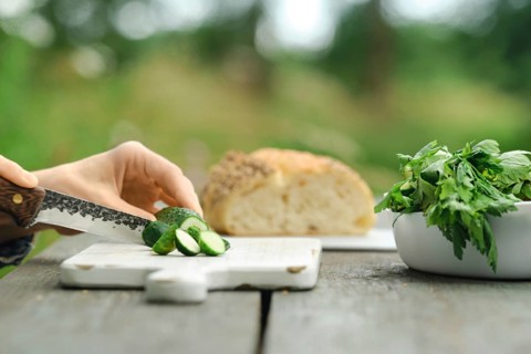 Person slices cucumbers on wooden cutting board. Fresh bread sits in background. Green herbs are
