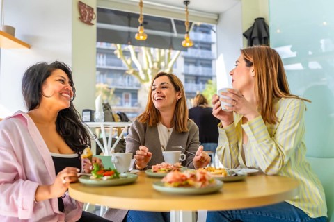 Three diverse women sitting at a table in a cafe, laughing and sharing a meal with drinks,