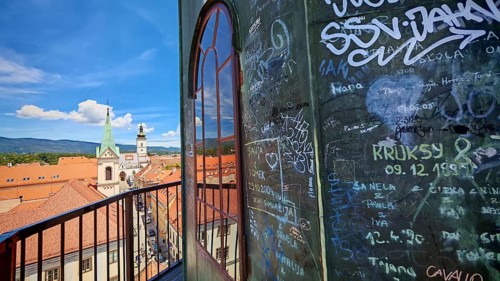 Scene of a tower with graffiti and a view from the Lotrscak Tower of a city with a dominant church,