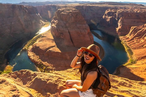 Young woman smiling, wearing a hat and sunglasses, sitting on a rocky cliff overlooking the iconic