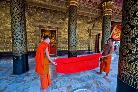 Buddhist monks and novices put together an orange monk's robe, sit in front of a large Buddha
