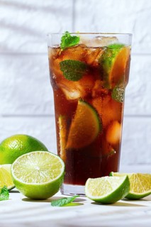 A glass filled with a cola beverage is surrounded by lime slices and fresh mint leaves. Whole limes