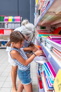 Grandmother and granddaughter actively selecting notebooks and stationery items from a store shelf.