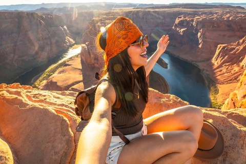 Young woman smiling and raising an arm, taking a selfie while sitting on a rock ledge at horseshoe